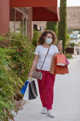 Photo of a young woman in mask shopping in the city while checking her mobile phone