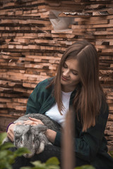 A girl in a green shirt is sitting in the garden with a light gray rabbit on her lap, her head cocked to one side.