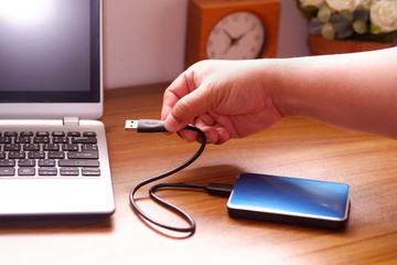 Man's hand was connecting an external portable hard disk usb 3.0 cable to laptop computer on desk,...