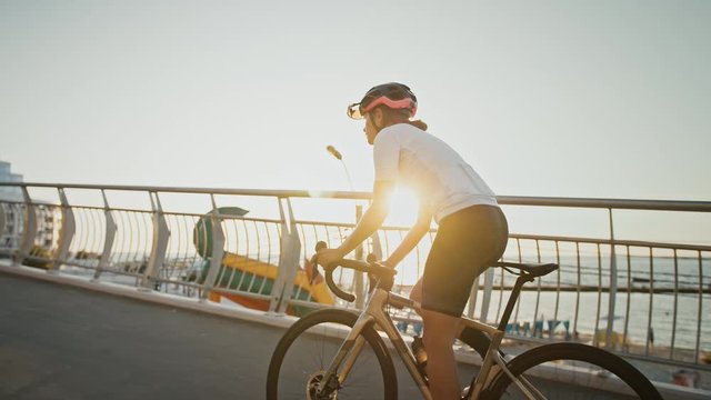 Female in sportswear and protective helmet is riding a sports bike uphill along bridge, near a sea coast. Close up, cinematic