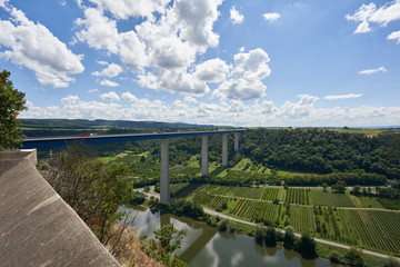 Moselle valley bridge made of blue steel and gray pillars leads over the river, green surroundings, blue sky with gray and white clouds. Germany.