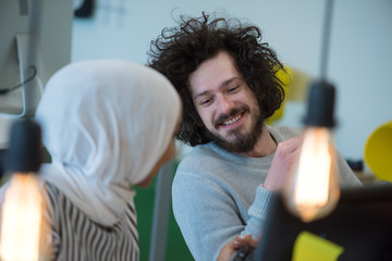 Smiling african muslim business woman talking to male colleague at work sharing office desk with desktops, friendly multiracial coworkers interns having pleasant fun conversation at workplace together