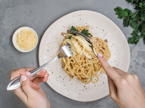 Women Eating A Plate Of Homemade Carbonara With Parsley, Crisp Bacon, Cheese And Garlic Garnished On Top At Home. Hands Twirl Spoon Fork To Roll Up Spaghetti As Italian Etiquette