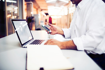 Skilled businessman synchronizes the mobile device to a portable laptop computer by using the application. Male freelancer sending e-mail on cell phone while standing in modern office interior