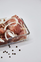Marinated fresh meat cut into pieces, lies in a glass dish form on a white background. Sliced onions on top of the meat and black pepper peas. vertical photo