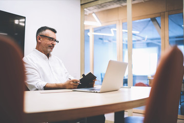 Male skilled jurist writing notes after consultancy with client. Mature editor creating new magazine article by using notebook and laptop computer. Experienced manager developing work-sharing plan