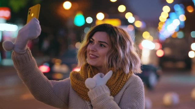 Portrait happy woman takes selfie on yellow phone, smiling happily while walking in warm knitted scarf, sweater in bokeh evening city in winter of Christmas lights. Lifestyle. Emotions. Holiday