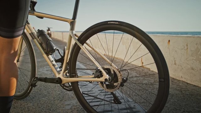 Unknown female in cycling shoes and sportswear is coming to concrete parapet, taking her bicycle, sitting down on it and riding along sea embankment
