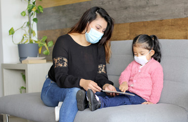 Latin mom with 2-year-old girl with protective face masks sitting in living room reading book, new...
