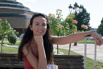 A smiling woman sitting on the street with a suitcase.