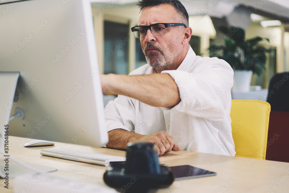 Aufkleber concentrated serious man in eye wear with black frame sitting at desktop with modern computer and wo