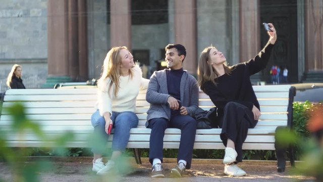 Three friends take selfies on city bench. Media. Friends take shared selfie sitting on bench while relaxing in city park. Shared selfie with three friends