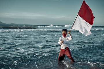 SchoolBoy in face mask Waving Red and White Flag on a Beach, commemorate Indonesia's independence day