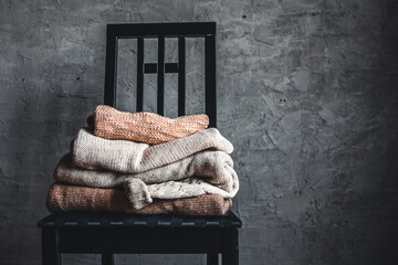 a stack of knitted warm cozy sweaters , on a chair by the gray wall. autumn, winter concept.
