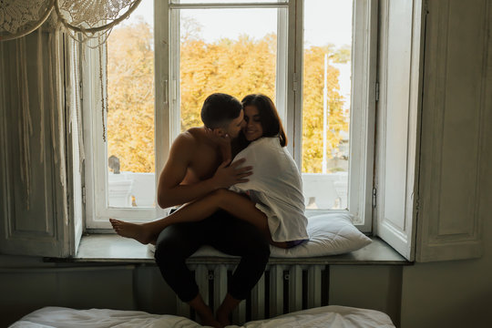 Young happy couple of man and woman at morning, sitting on windowsill hugging and kissing. 
