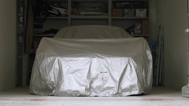 A sports car is parked under an awning in a bright home garage. Footage. Opens the gates of a white garage, inside a car closed with a cloth and shelves with a tool in the background
