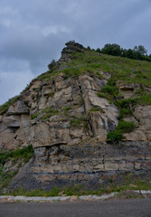 the roadbed is uneven, winding, passes through mountains and cliffs. Clouds are dark, low, with rain. A thick white fog is spreading along the road.