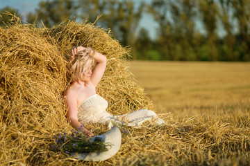 A woman in a white dress in a haystack at sunset. An evening of peace and quiet. A mother of three boys, a wonderful figure. Light hair, light clothing, spikelets in the field. Belarus.