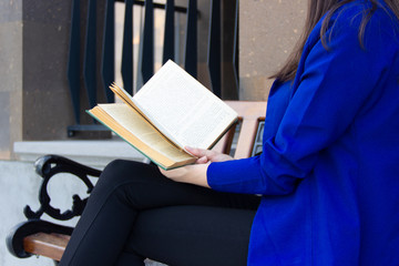 A woman in a blue jacket is reading a book sitting on a bench