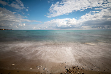 Seawaves splashing onto the sandy coast with pebbles and blue cloudy sky.