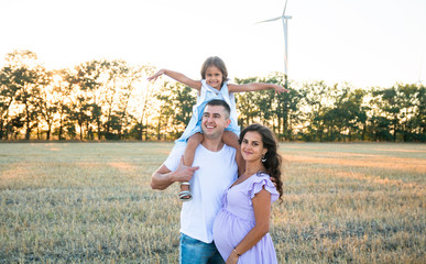 happy young husband, pregnant wife and a little girl on the father's shoulders on the field, wind turbines on the background