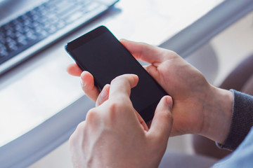 photo of the hands of a young man who is dressed in a gray sweatshirt and holding a black phone. Against the background of a white table with a laptop on it