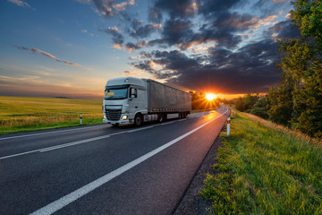 White truck driving on the asphalt road in rural landscape in the rays of the sunset with dark storm cloud
