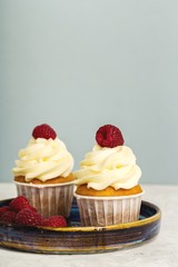 Two cupcakes with cream cap and raspberries on blue plate, blue paper background. Space for text, vertical format.