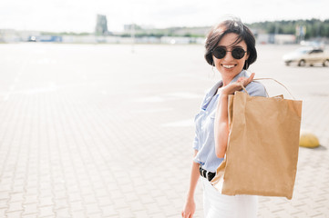 Shopping time. Trendy woman is happy with shopping. A young caucasian smiling woman in a stylish wear and sunglasses holds paper bags in hand smiling and looks at the camera