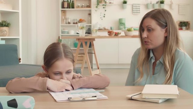 Lockdown of irritated Caucasian woman is sitting at desk in living room, checking her daughter home task and scolding her because of mistakes. Girl is upset