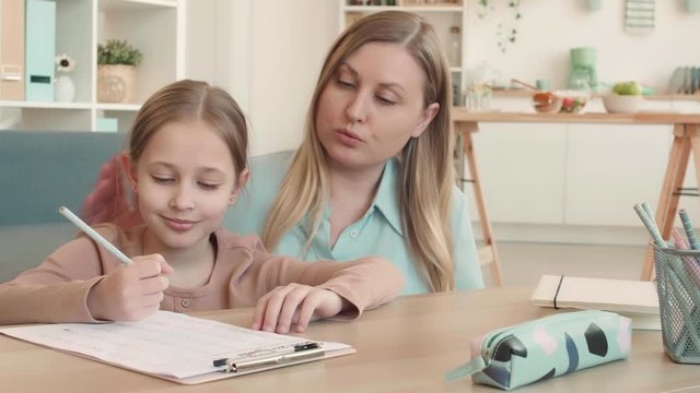 Medium shot of attractive Caucasian woman and schoolgirl with blond hair are sitting at desk in living room and doing math homework together