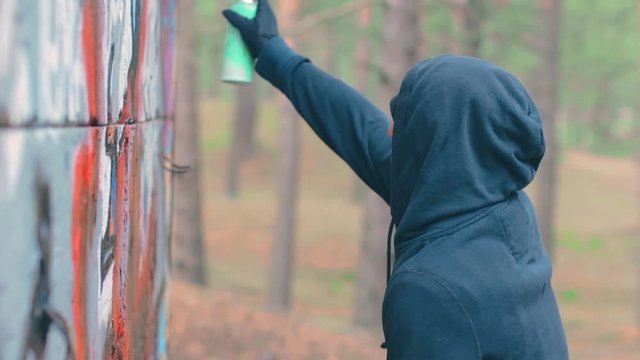 Youthful Guy Vandalizes the Wall Using a Spray Paint Can. Hooligan in Casual Clothing is Drawing Graffiti Outdoors. Youth Movement Crime Related with Street Art. Middle Close-Up View, Dynamic Shot