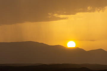 Mountainous landscape at sunset with a distant curtain of rain on the horizon and the sun disk hiding