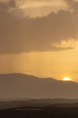 Mountainous landscape at sunset with a distant curtain of rain on the horizon and the sun disk hiding