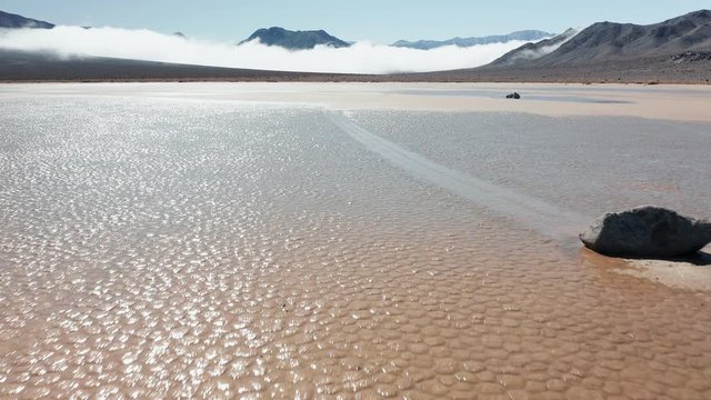 Cinematic 4K aerial of moving stones on sunny morning, with mountains and clouds on background. Close up of black rock on the wet cracked surface of Death Valley desert. Epic race track playa park