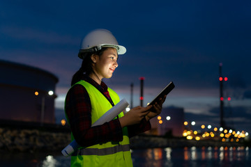 Asian woman petrochemical engineer working at night with digital tablet Inside oil and gas refinery...