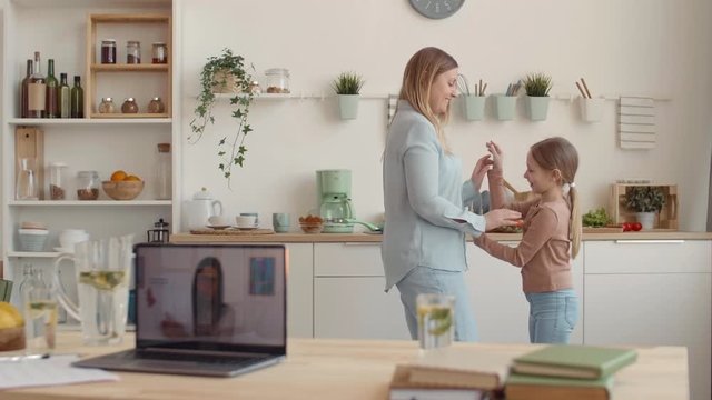 Lockdown of laptop with video lesson on its screen standing on kitchen table while Caucasian blond-haired woman and her daughter having fun on background
