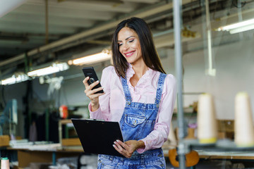 Happy young entrepreneur smiling at work while holding mobile phone - Female manager or worker making video call or texting in factory - success achievement happiness and independence equality concept