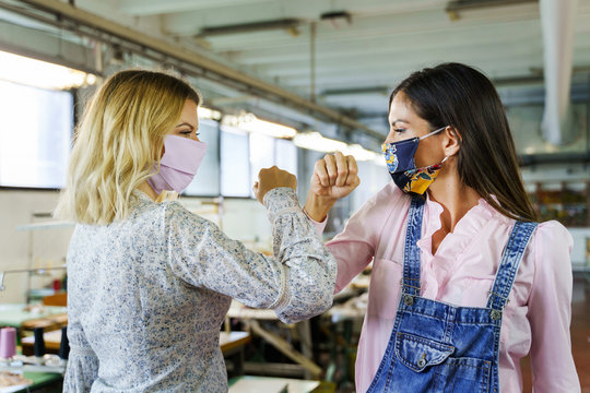 Two women greeting at work during covid-19 pandemic - Female friends workers elbow bump wearing protective masks in factory - front view in bright day - Real people new normal social distance concept