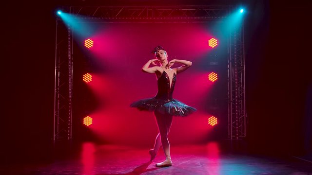Graceful ballerina in a chic image of a black swan. Young beautiful girl in a black tutu with red sequins and a crown. Shot in a dark studio with smoke and red neon lighting. Slow motion.