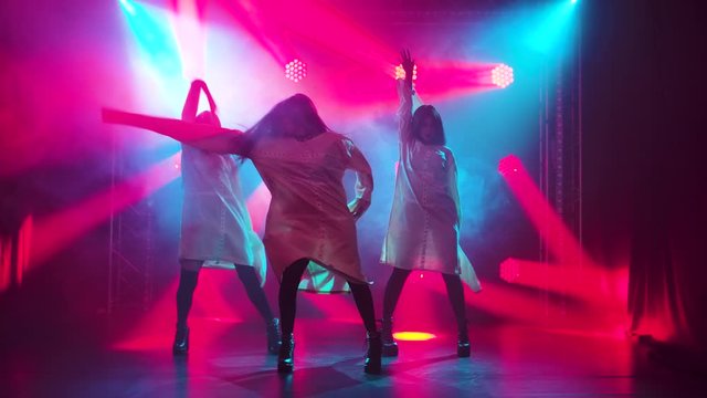 Three charming girls in white straitjacket are dancing on the stage. Silhouettes in a dark studio with smoke and neon lighting. Red neon light effects. Dance show. Slow motion.