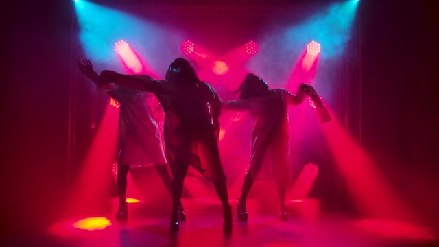 Three charming girls in white straitjacket are dancing on the stage. Silhouettes in a dark studio with smoke and neon lighting. Red neon light effects. Dance show. Slow motion.
