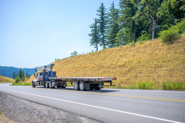 Big rig semi truck with crane application on the frame transporting empty flat bed semi trailer running on the winding road with yellow summer hills