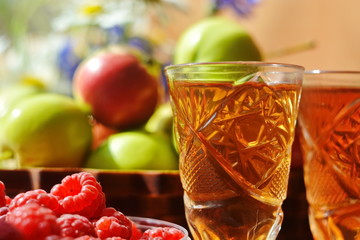 Shot of berry drink, wine or cognac next to glass of fresh ripe raspberries close up on blurred background of bunch of flowers and basket of apples