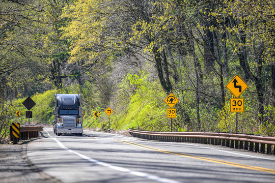 Classic Black Big Rig Semi Truck With Semi Trailer Climbing Up Hill On The Forest Winding Summer Road