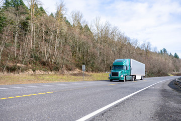 Green bonnet big rig semi truck transporting cargo in refrigerator semi trailer driving on the road with forest on the hill side