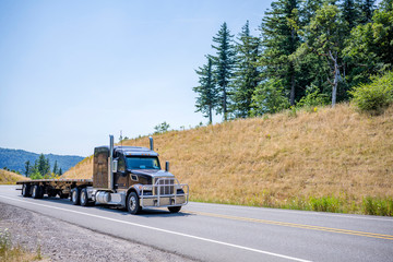 Fototapeta na wymiar Powerful big rig black semi truck with grille guard going up hill with empty flat bed semi trailer in gorgeous Columbia Gorge area