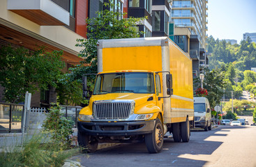 Yellow big rig semi truck delivering cargo in box truck standing on the street parking spot with multilevel buildings