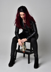 Fototapeta premium Full length portrait of girl with red hair wearing black leather jacket, pants and boots. Sitting pose on a chair holding a gun, isolated against a grey studio background.