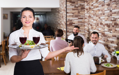 Pretty female waiter greeting customers at table in restaurant